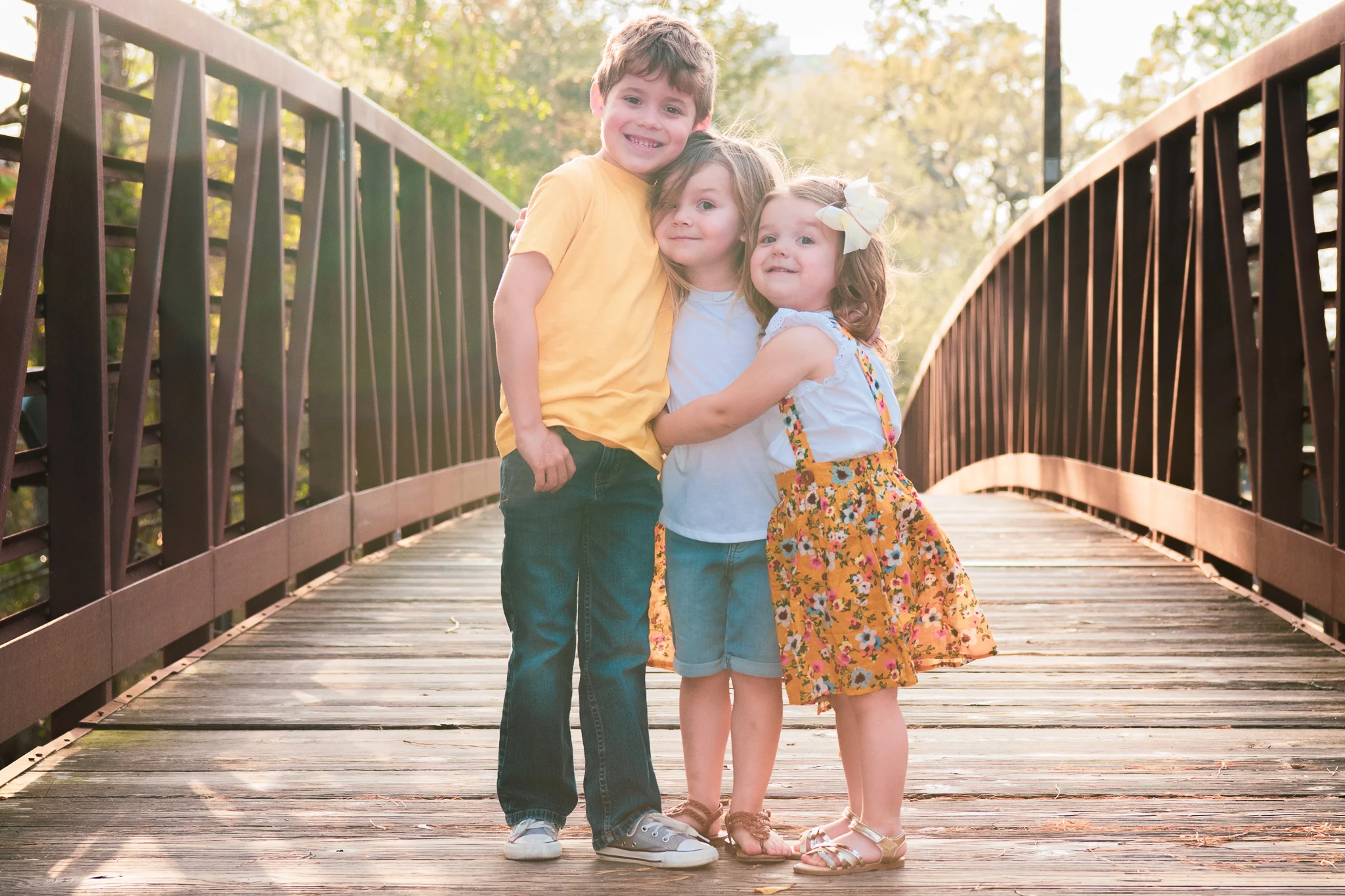Three siblings embracing on a sunlit bridge.